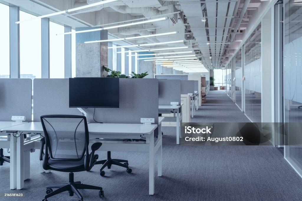 The modern open-plan office with a clean, organized layout. The workspace features rows of individual desks equipped with computers and ergonomic office chairs, separated by privacy screens.